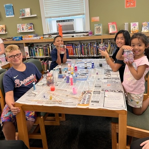 Four children sitting at a table in the library, displaying their homemade Galaxy Jars for Tween Craft Club 