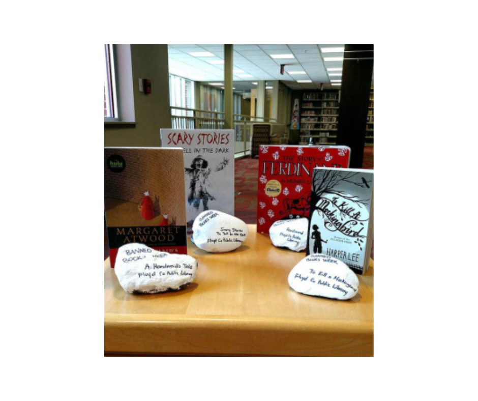 copies of banned books displayed on a table in a library with white rocks in front of the books.  On each rock is painted the name and author of the book that it is in front of on the table.