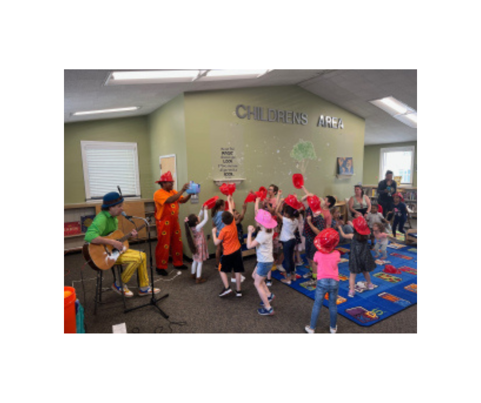 image of the library childrens' room with children dancing, a man seated and playing a guitar, and a man standing and dancing