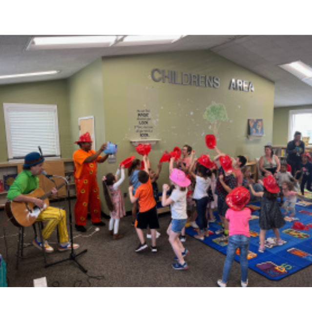 image of the library childrens' room with children dancing, a man seated and playing a guitar, and a man standing and dancing