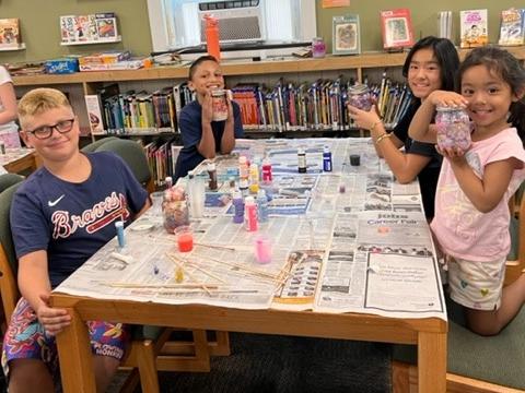 Four children sitting at a table in the library, displaying their homemade Galaxy Jars for Tween Craft Club 
